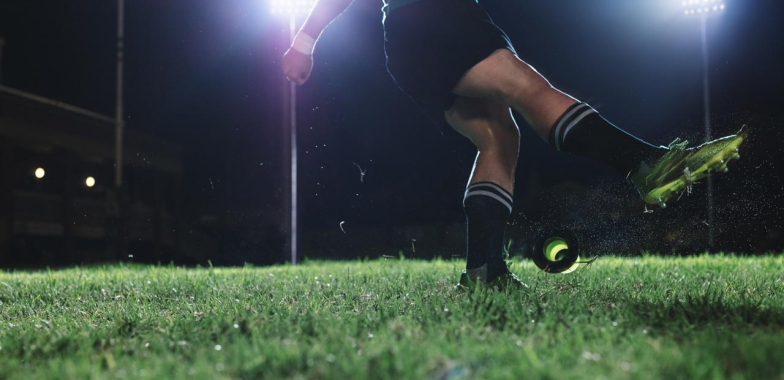 rugby player kicking a ball on pitch at night with spotlight on