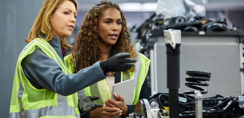 Two women in hi vis vests in a factory discussing a machine