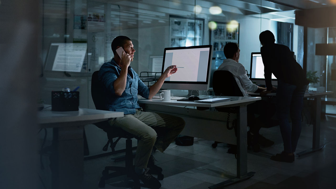 Man sitting at desk in an office, on the phone and pointing at his computer screen